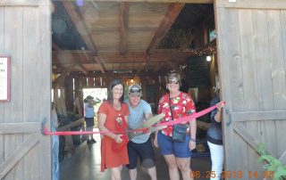 Three individuals smiling at the camera during a ribbon-cutting ceremony at a wooden entrance.